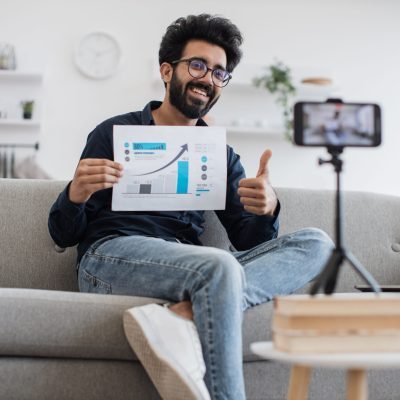 Young arabian financier with black beard filming himself on cell phone which fixed on tripod and showing financial statistics to subscribers in living room thumb up. Concept of technology and blogging